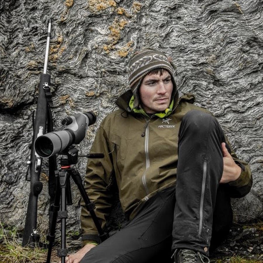 Logan Evans against folded grey rock with spotting scope and rifle.